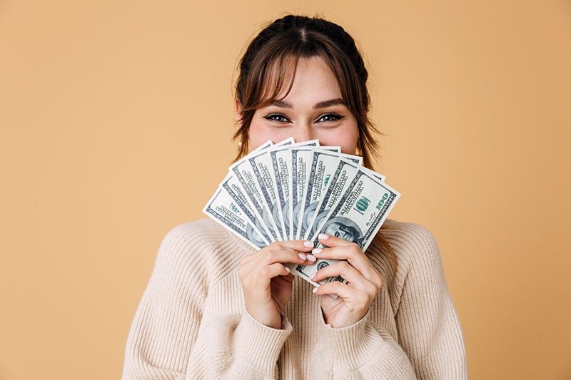 Young woman wearing sweater standing against beige background, showing money fanned out in her hands - a common question is which destination is more expensive between Naples and Marco Island